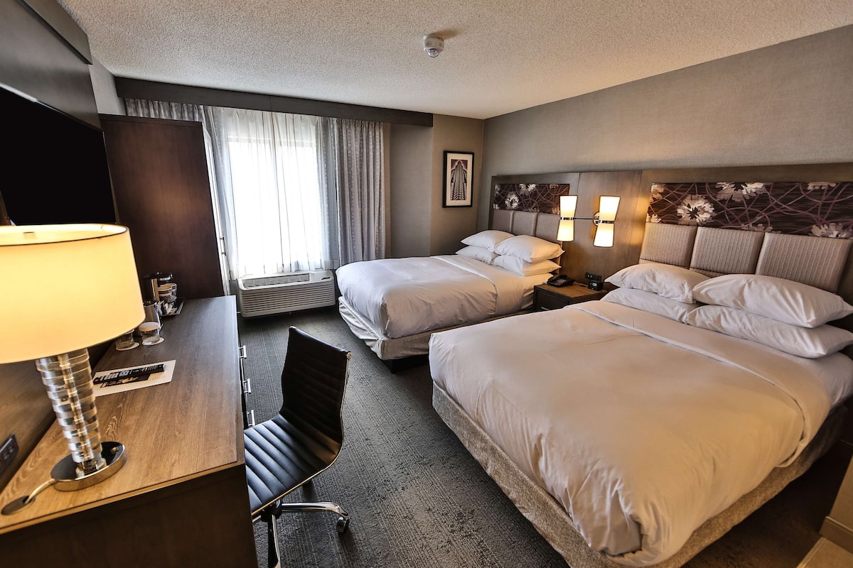 A hotel room with two beds, a desk with a lamp, a chair, and a window with curtains.