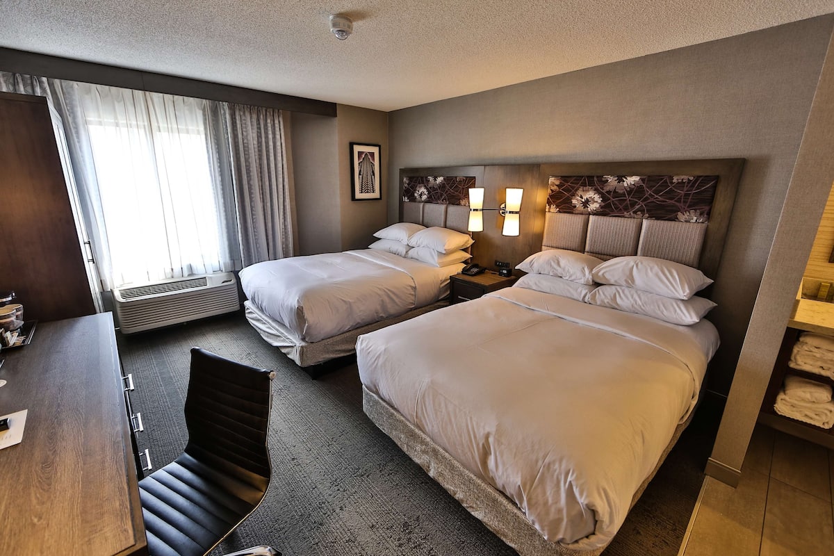 A hotel room with two beds, a desk, a chair, a heater, and a large window with curtains.