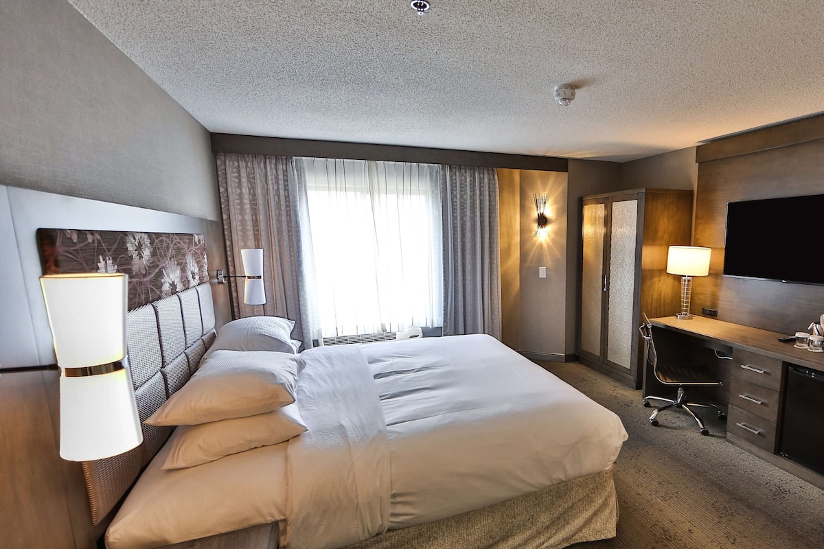A hotel room with a large bed, a desk with a chair, a television, and a wall-mounted lamp.
