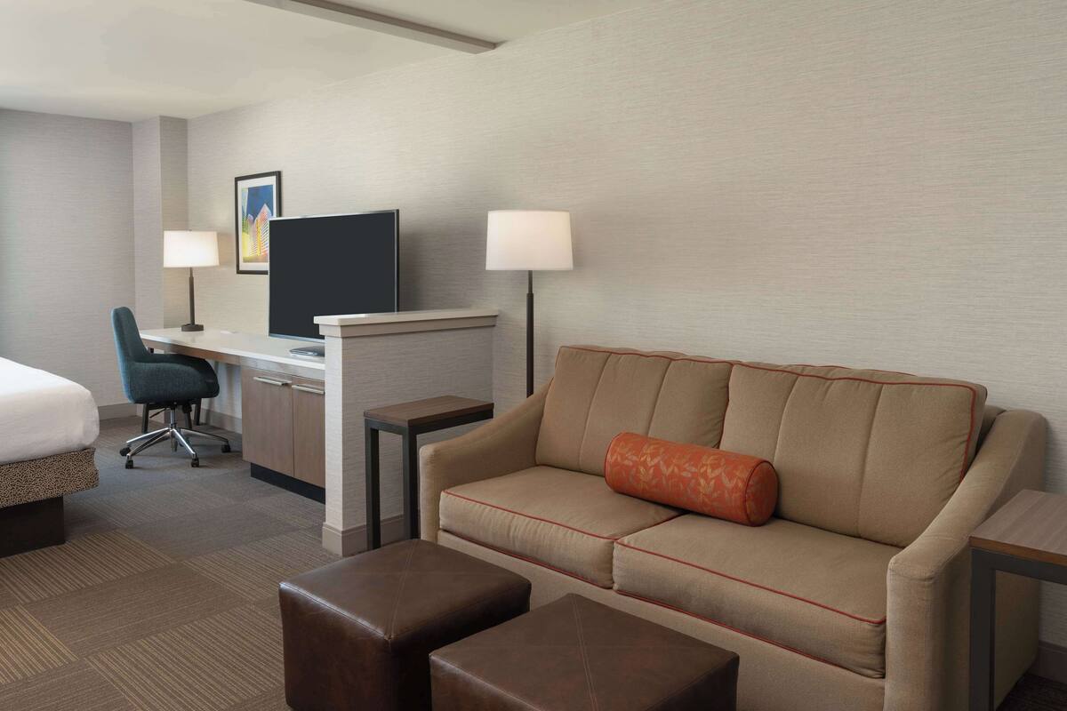 A hotel room with a bed, a desk with a computer, a sofa with a cushion, and two ottomans.