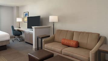 A hotel room with a bed, a desk with a computer, a sofa with a cushion, and two ottomans.