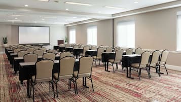 Un salón de conferencias con filas de mesas y sillas, una pantalla de proyector y ventanas amplias.