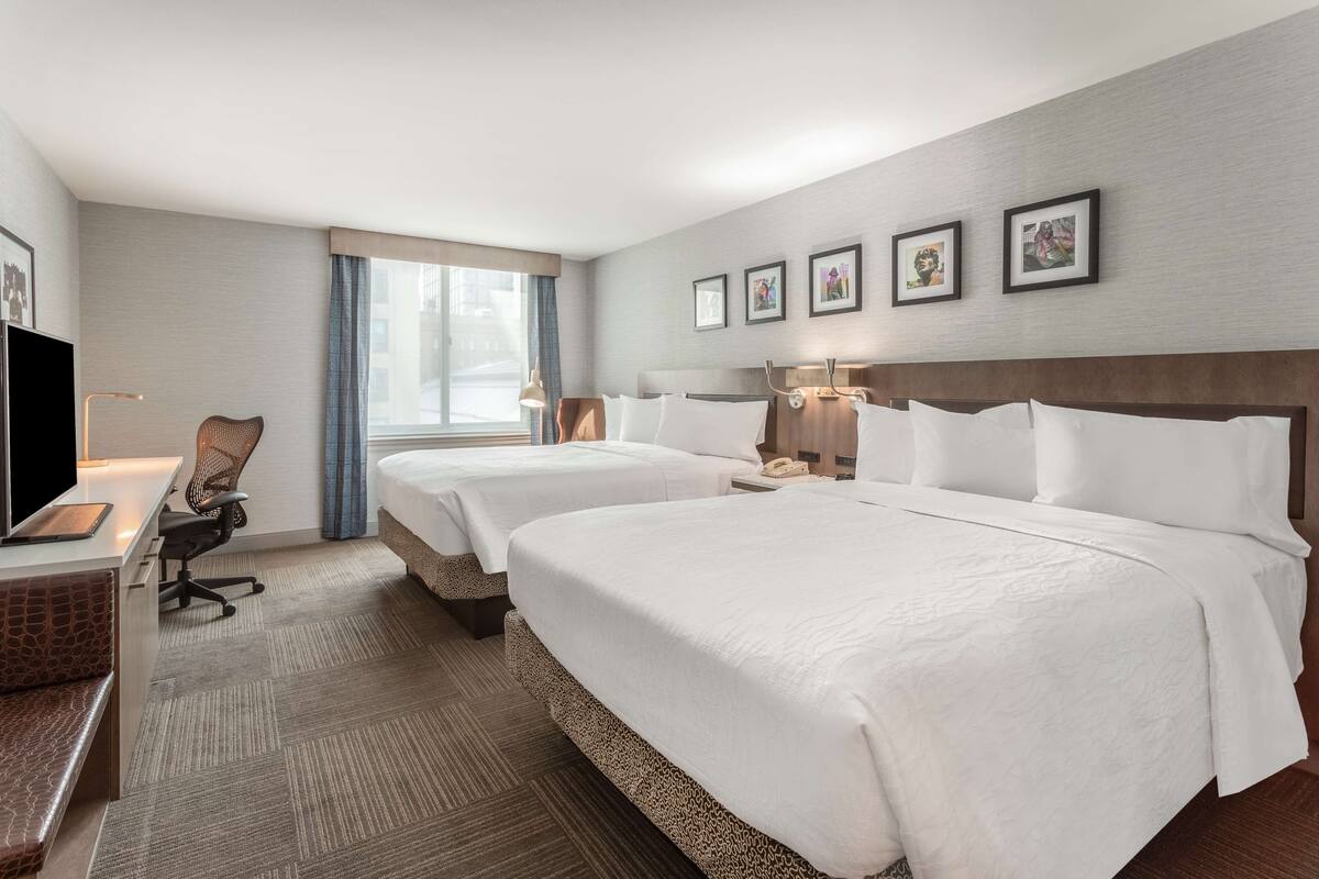 A hotel room with two beds, a desk with a computer, a chair, a TV, and framed pictures on the wall.