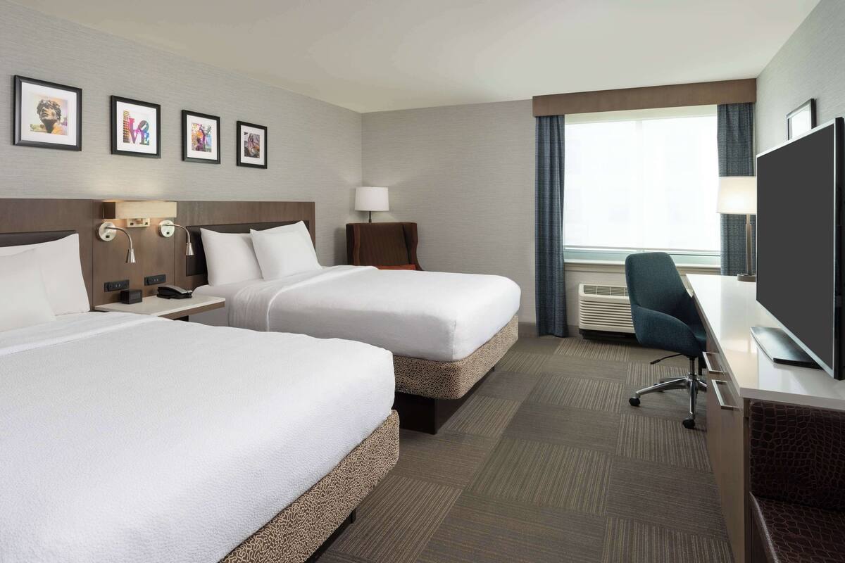 A hotel room with two beds, a desk, a TV, and a window with curtains.