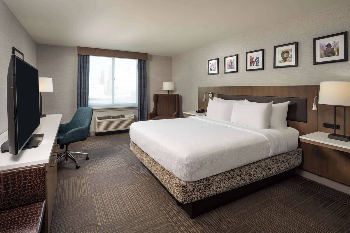 A hotel room with a large bed, a desk with a computer, a chair, a window with curtains, and framed pictures on the wall.