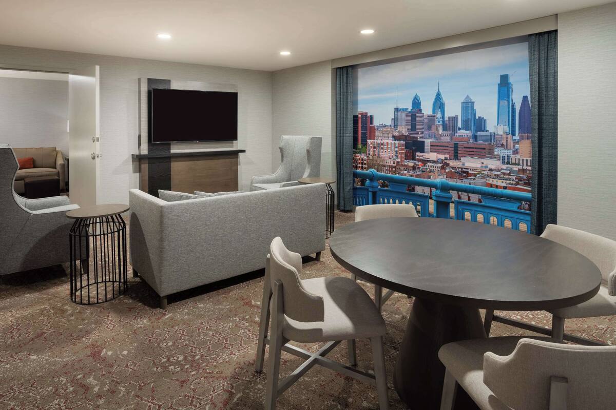 A hotel room with a cityscape view, a flat-screen TV, a sofa, a dining table with chairs, and a patterned carpet.