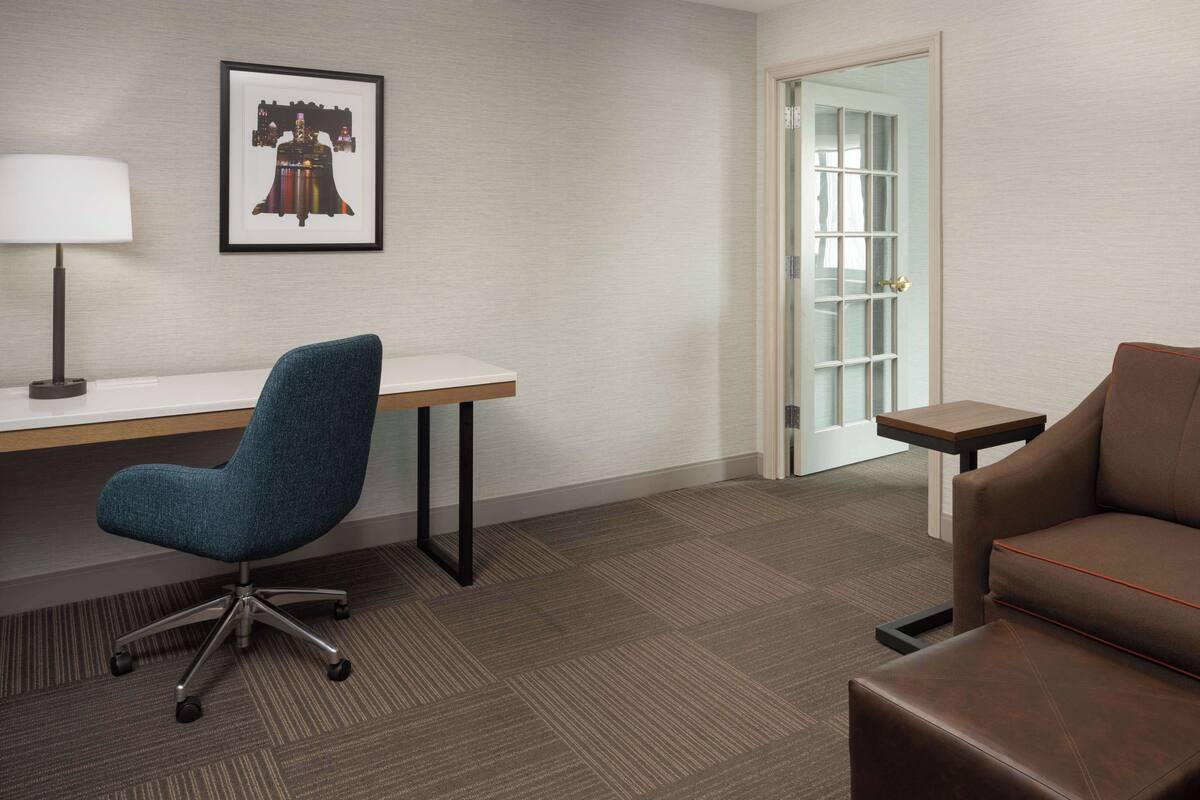 A hotel room with a desk, a blue office chair, a brown sofa, a framed artwork on the wall, and a door leading to another room.