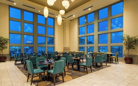 A spacious hotel lobby with large windows, multiple dining tables, and green upholstered chairs.