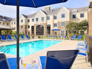 A swimming pool with lounge chairs and umbrellas, surrounded by a building with stone and stucco exterior. - Sonesta ES Suites Charlotte Arrowood (Charlotte)