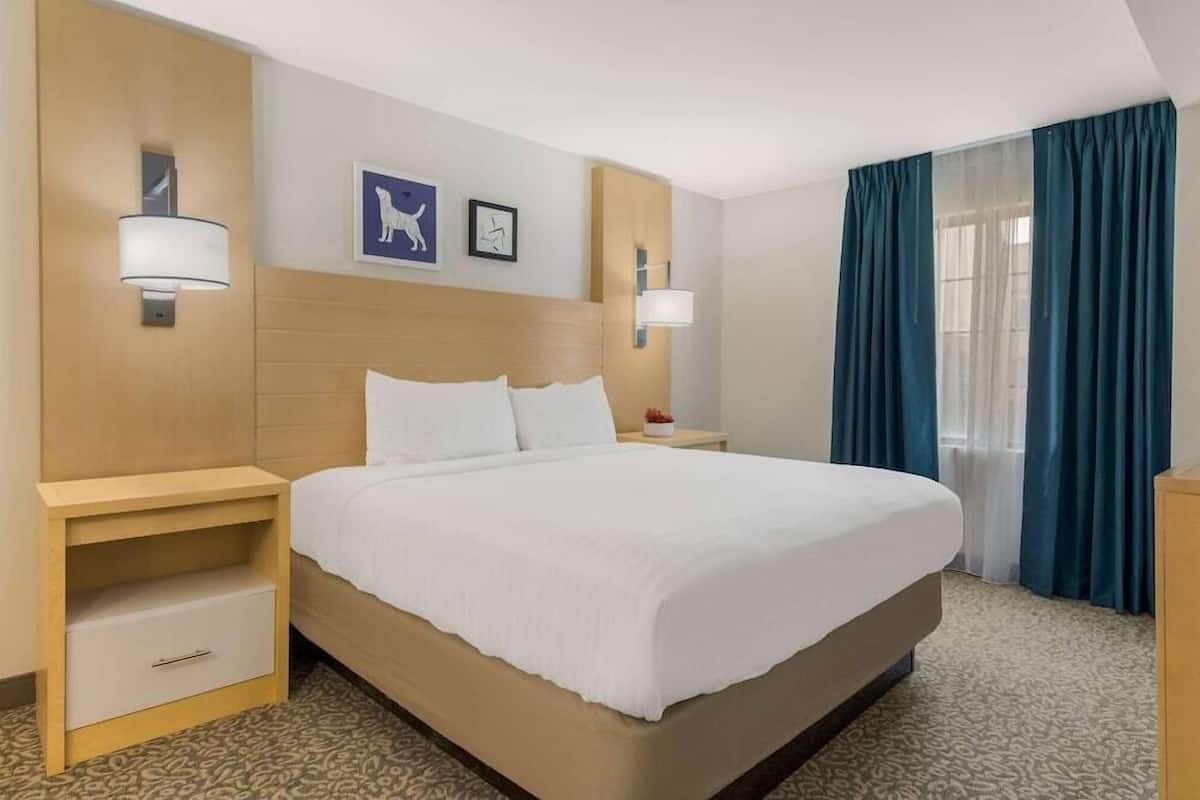 Habitación de hotel con una cama grande, dos mesitas de noche, una lámpara de pared, un cuadro enmarcado y una ventana con cortinas.
