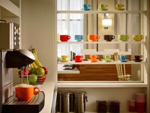 Une cuisine équipée d’une machine à café, d’un panier de fruits et d’une étagère où sont rangées des tasses et des soucoupes aux couleurs vives.