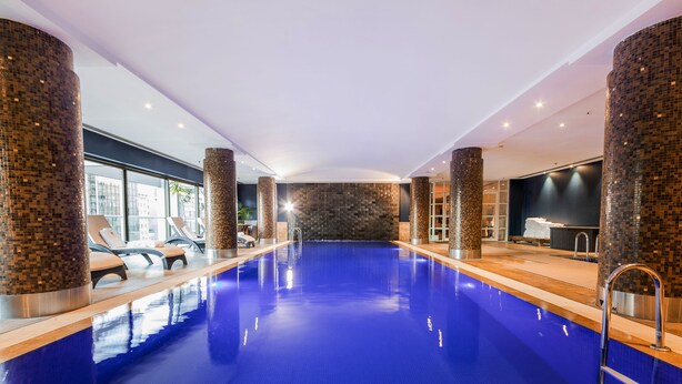 Una piscina interior moderna con columnas revestidas de mosaicos, sillas de descanso y vistas a la ciudad.