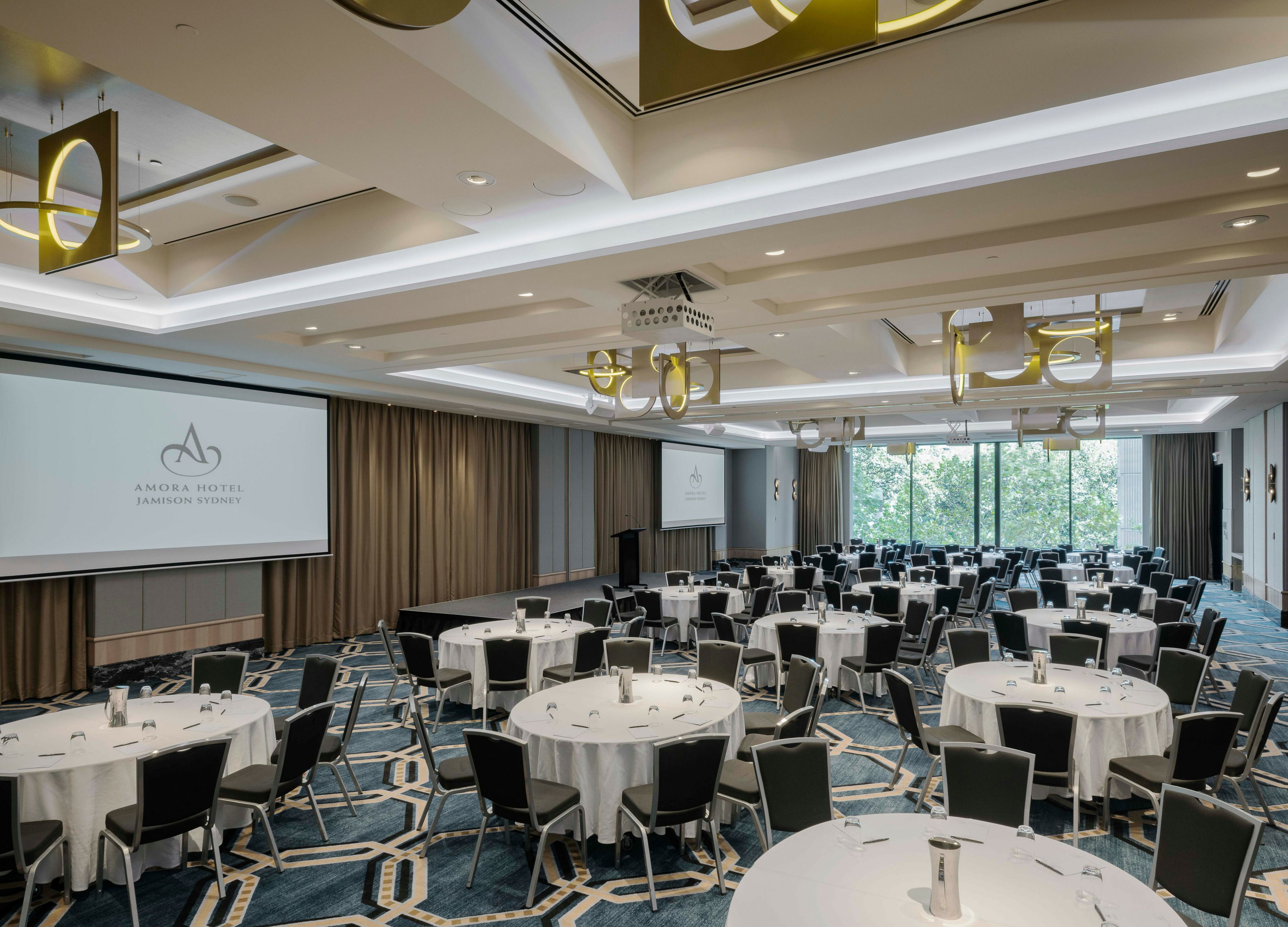 A conference room with multiple round tables, chairs, and a projection screen displaying the hotel name.