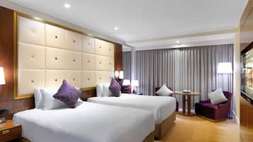 A hotel room with two beds, a large padded headboard, a small round table, and a seating area with a purple armchair and a sofa.