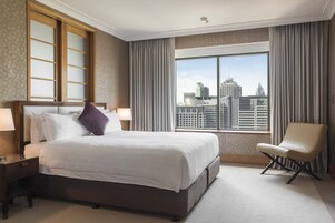 Habitación de hotel con una cama grande, una silla, una mesita de noche y una ventana con vista a la ciudad.