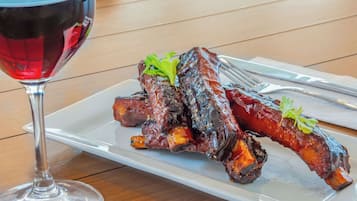 Prato de costelas de churrasco, decorado com salsa, acompanhado de um copo de vinho tinto, sobre uma mesa de madeira.