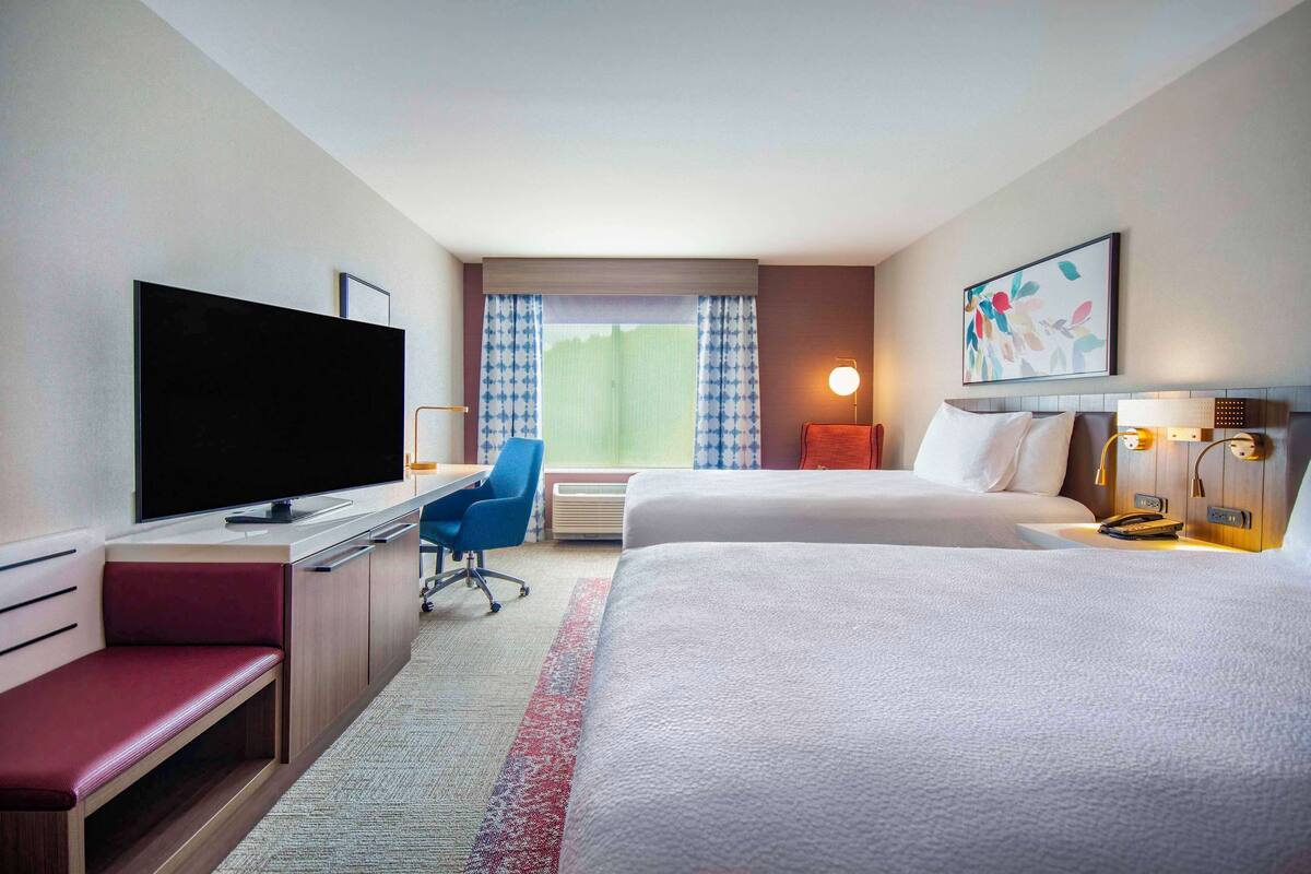 A hotel room with a large flat-screen TV, a desk, a blue office chair, two beds, a red bench, a window with curtains, and a painting on the wall.