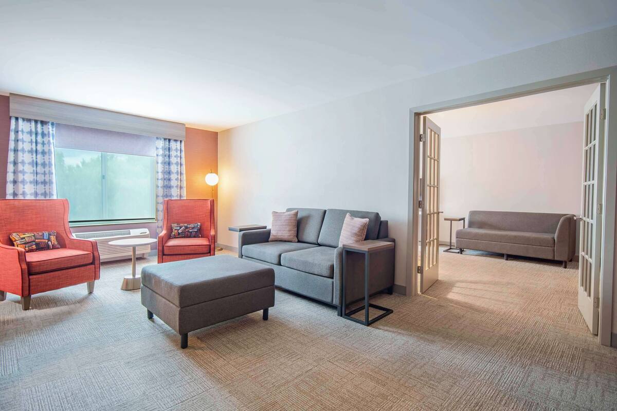 A modern living room with a grey sofa, red armchairs, and a grey ottoman. There is a large window with patterned curtains, a small side table, and a lamp.