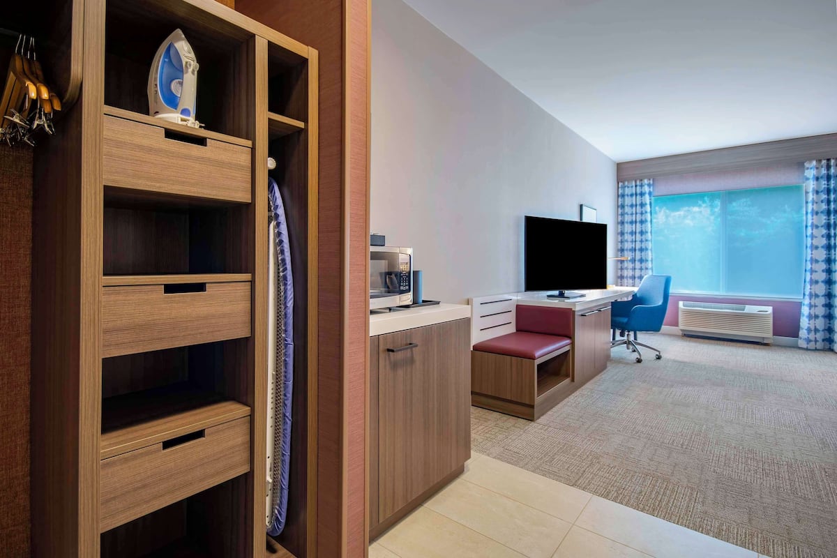 A hotel room with a wooden wardrobe, a desk with a computer, a TV, and a large window with curtains.