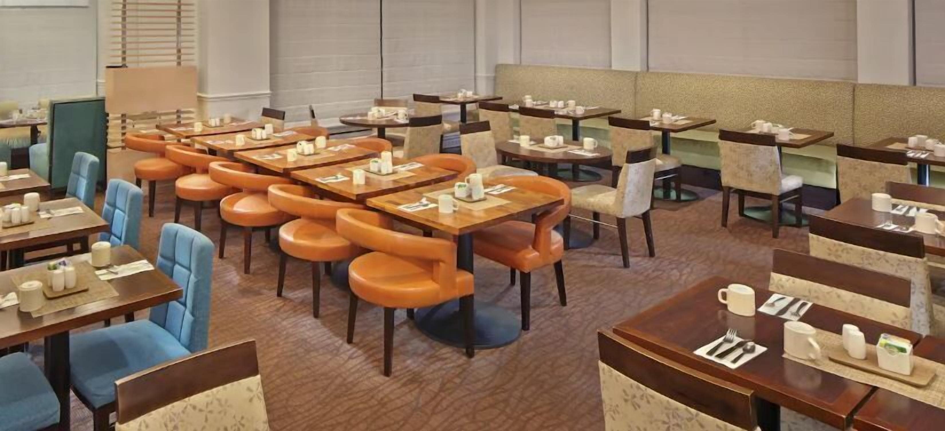 A hotel dining area with multiple tables and chairs, some with orange cushions.