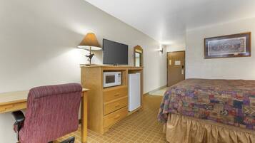 Una habitación de hotel con un escritorio de madera, una silla roja, una cama con colcha estampada, un televisor, un microondas y un refrigerador.