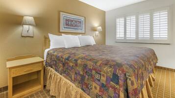 A hotel room with a bed, two bedside lamps, a nightstand, a patterned bedspread, a wall-mounted picture, and a window with blinds.