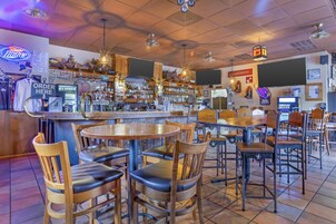 Una zona de bar con mesas y sillas de madera, una grifería de cerveza y varias botellas y vasos en estantes.