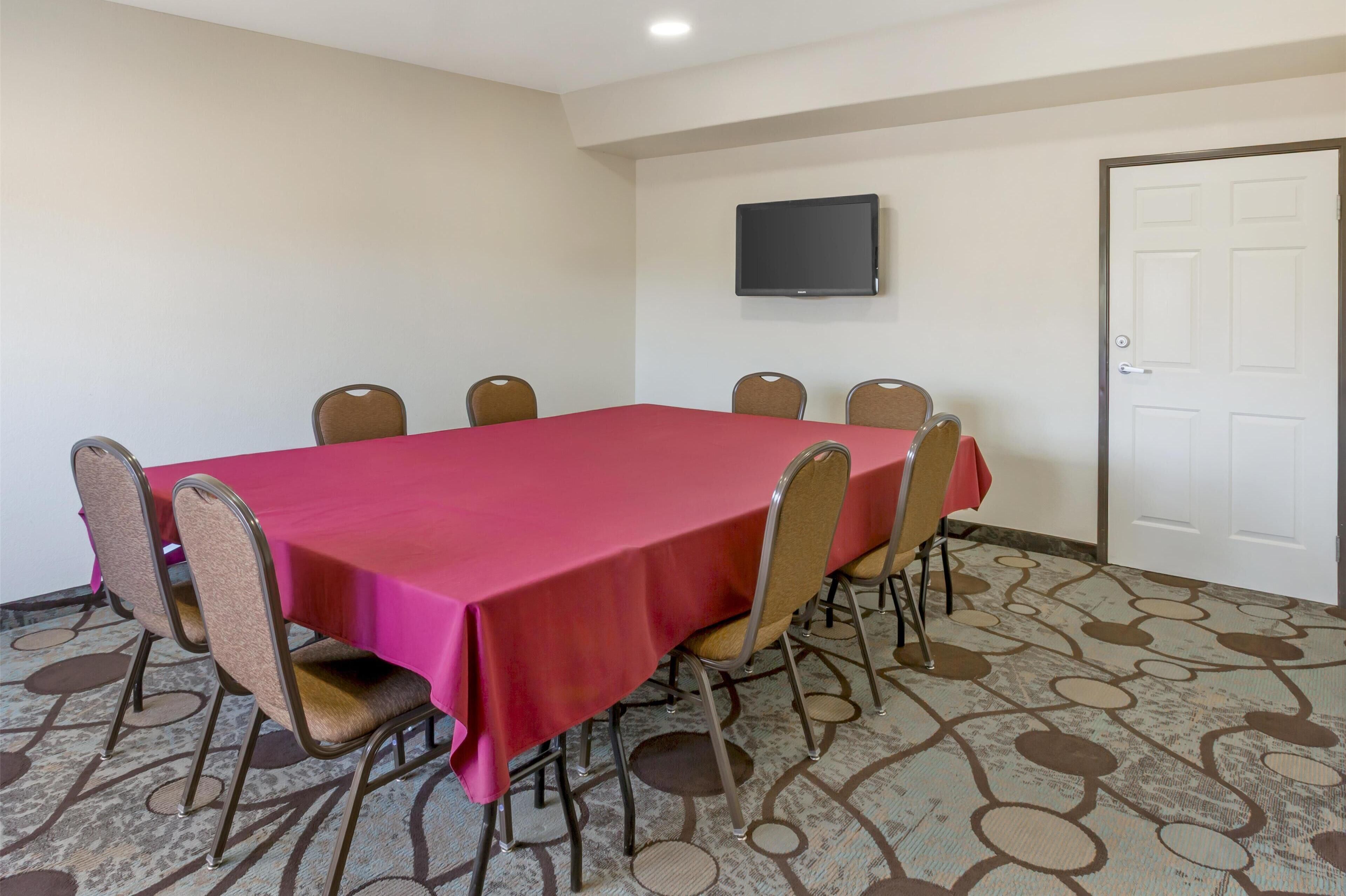 A conference room with a red table, beige chairs, a flat-screen TV on the wall, and a patterned carpet.