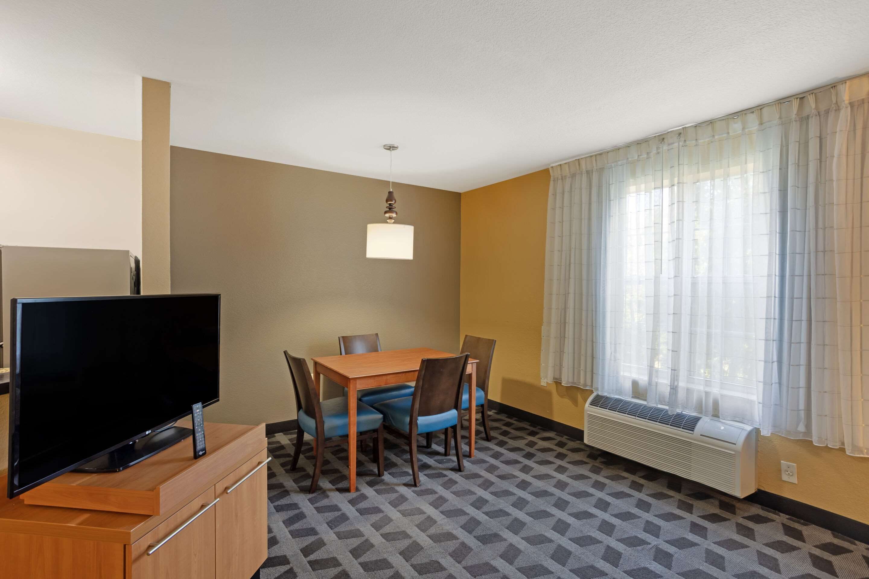 A hotel room with a flat-screen TV on a wooden cabinet, a dining table with chairs, a hanging light fixture, a window with sheer curtains, and a wall-mounted air conditioner.