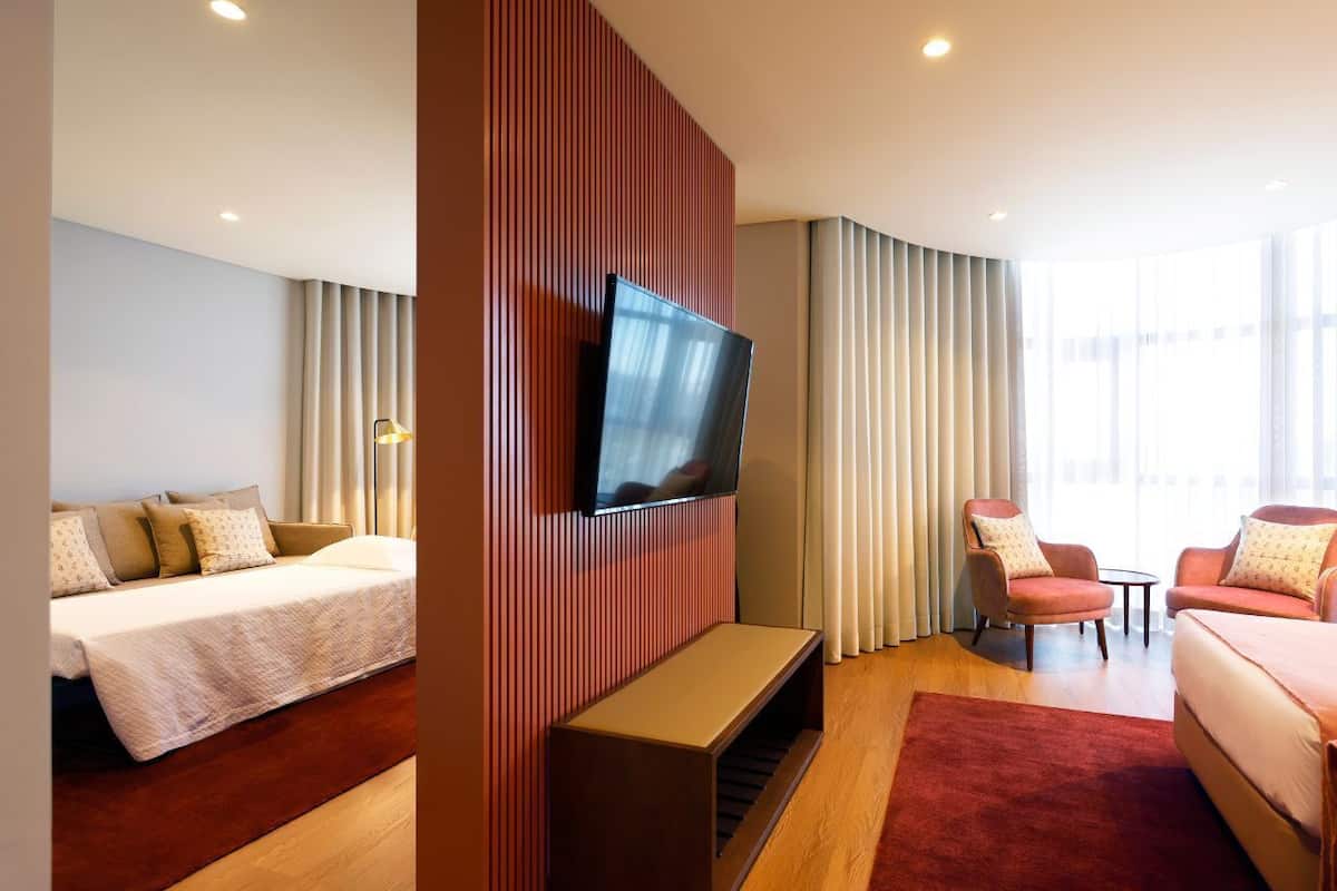 Moderni hotellihuone, jossa on sänky, oleskelualue, televisio ja suuret ikkunat.