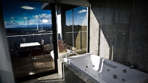 A modern bathroom with a large bathtub, a glass door leading to a balcony with a table and chairs, and a view of a cityscape. - Montebelo Viseu Congress Hotel & Spa (Viseu)