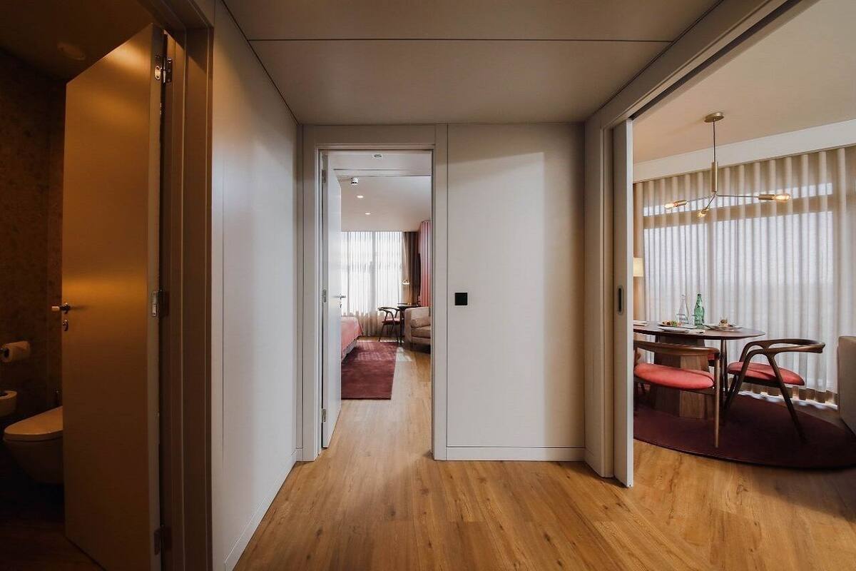 Moderni hotellihuone, jossa on puulattia, ruokailutila pyöreän pöydän ja tuolien kera sekä näkyvissä oleva kylpyhuone, jossa on wc.