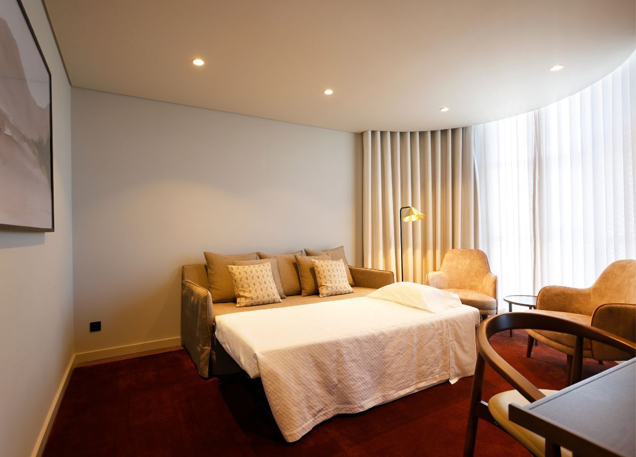 Une chambre d’hôtel avec un lit, deux fauteuils, une petite table et une fenêtre avec des rideaux.