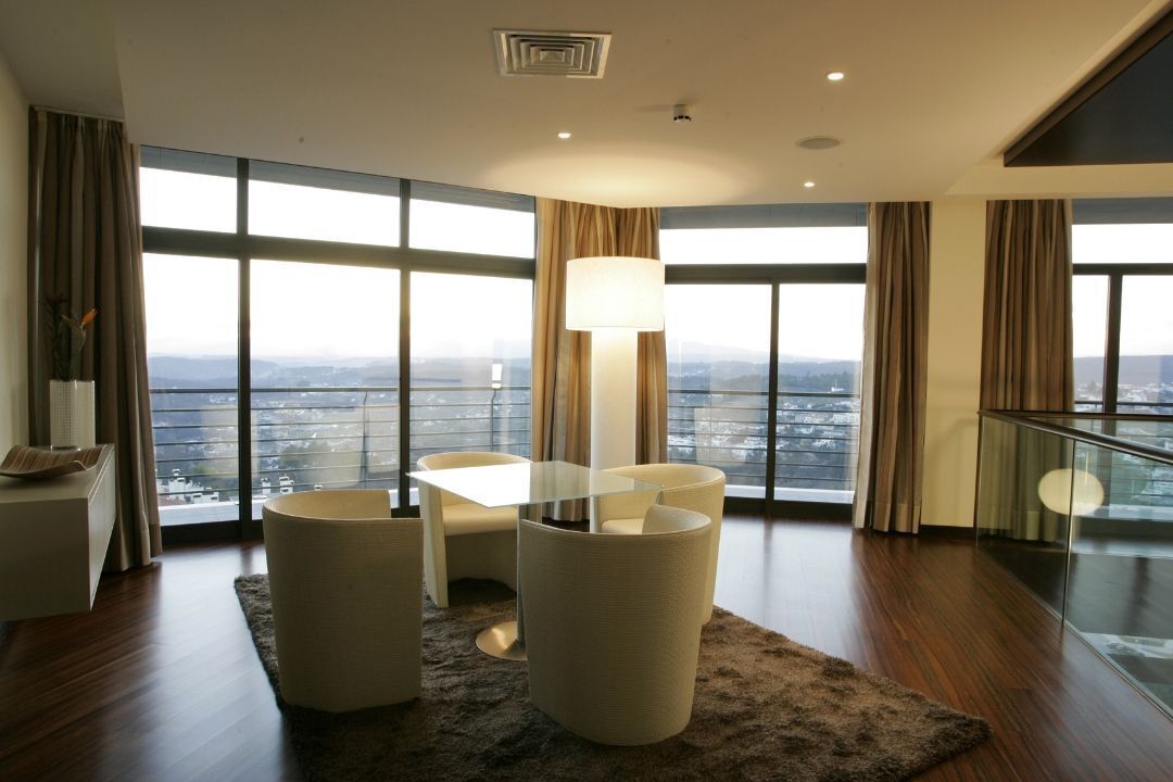 A modern living room with a glass table, a large floor lamp, and a view of the city through large windows.