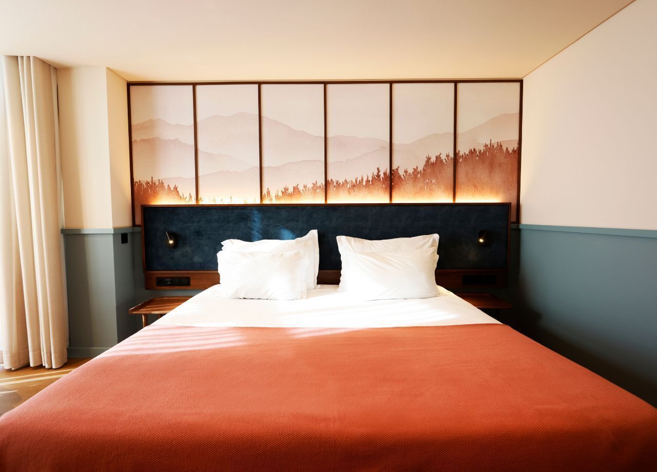 A hotel room with a large bed, a headboard with a cityscape artwork, and a nightstand with a lamp.