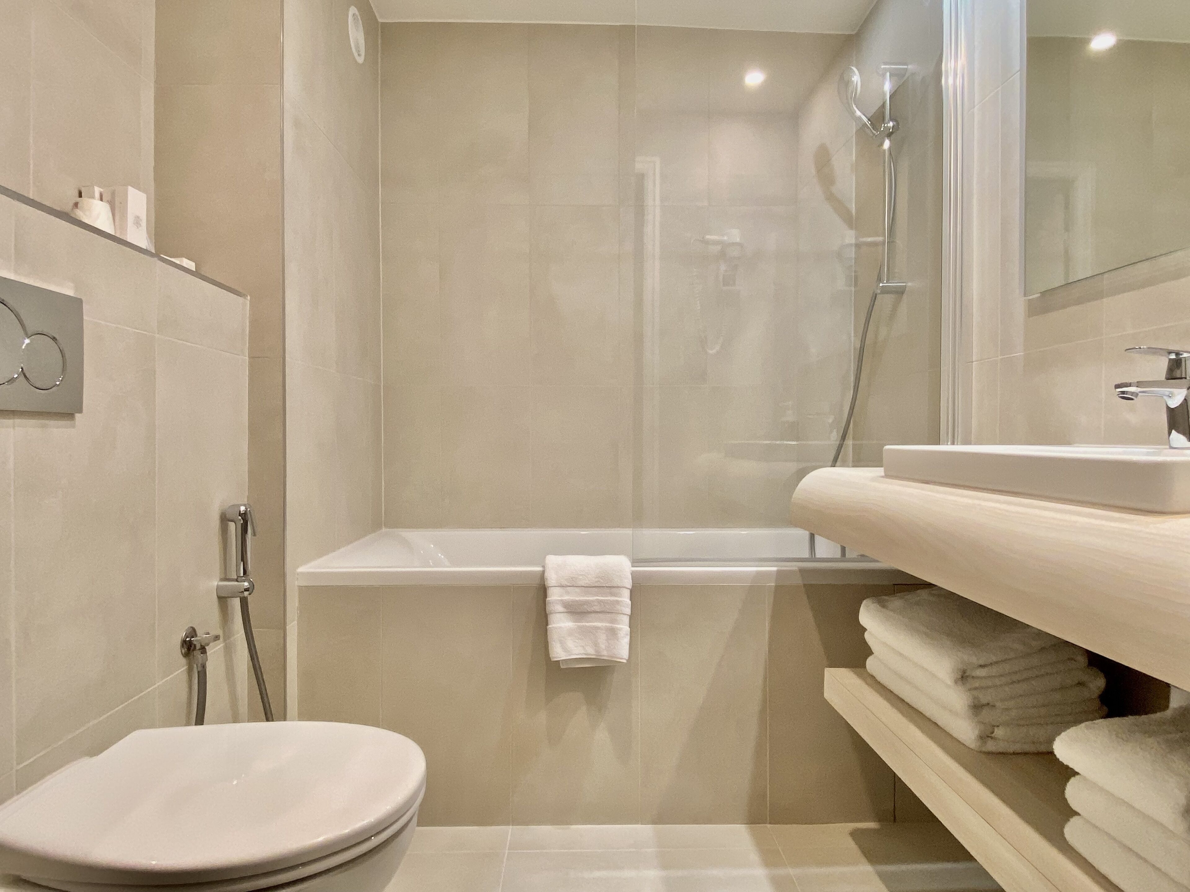 A modern bathroom with a toilet, bathtub, and shower. There is a towel rack and a shelf with folded towels.