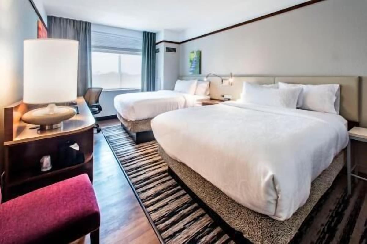 A hotel room with a large bed, a smaller bed, a desk with a lamp, a chair, and a window with curtains.