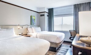 A hotel room with two beds, a desk with a lamp, a painting on the wall, and a window with curtains. - Hilton Garden Inn Minneapolis Airport Mall of America (Bloomington)
