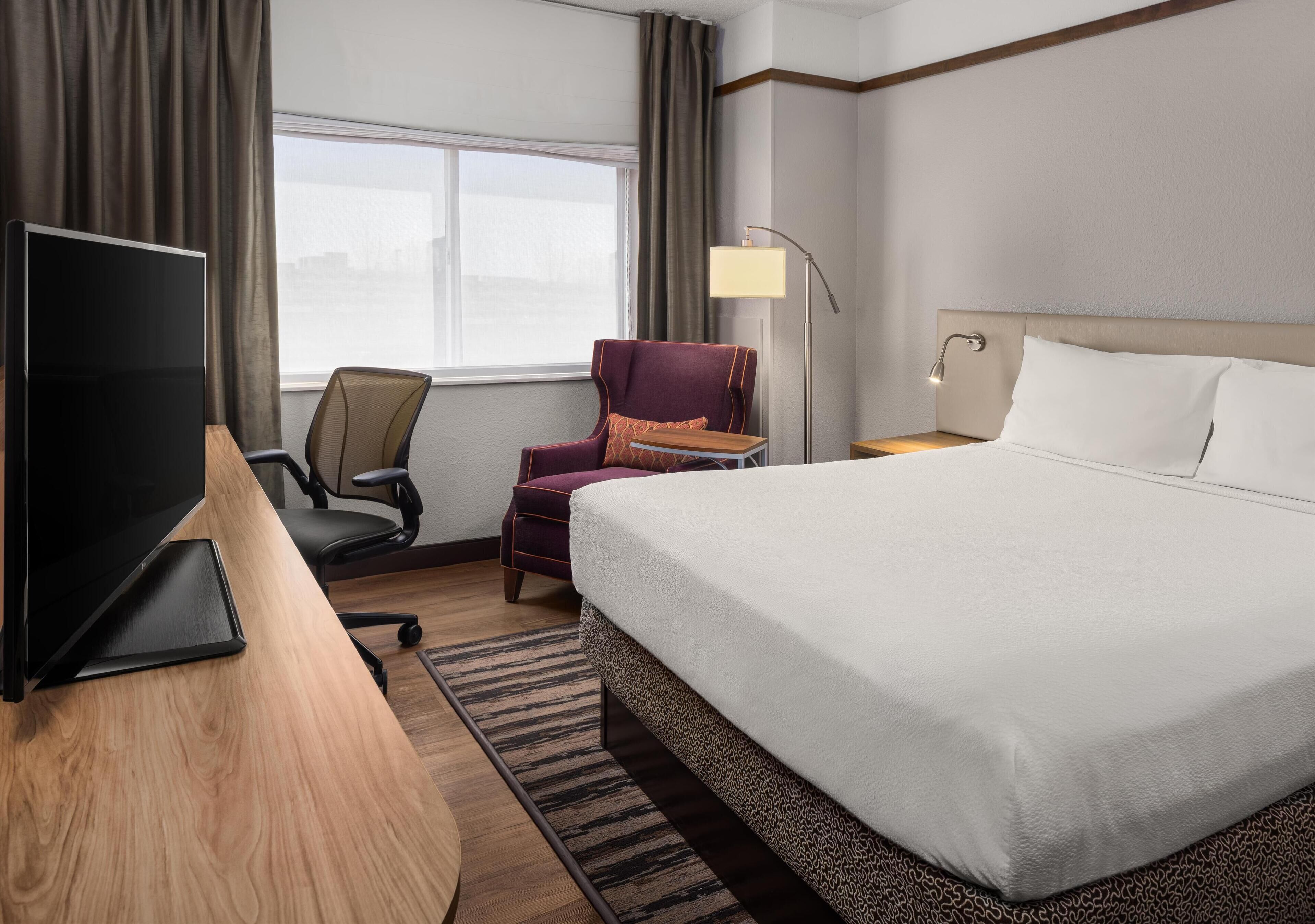 A hotel room with a large bed, a desk with a laptop, a chair, a lamp, and a view of the city through the window.