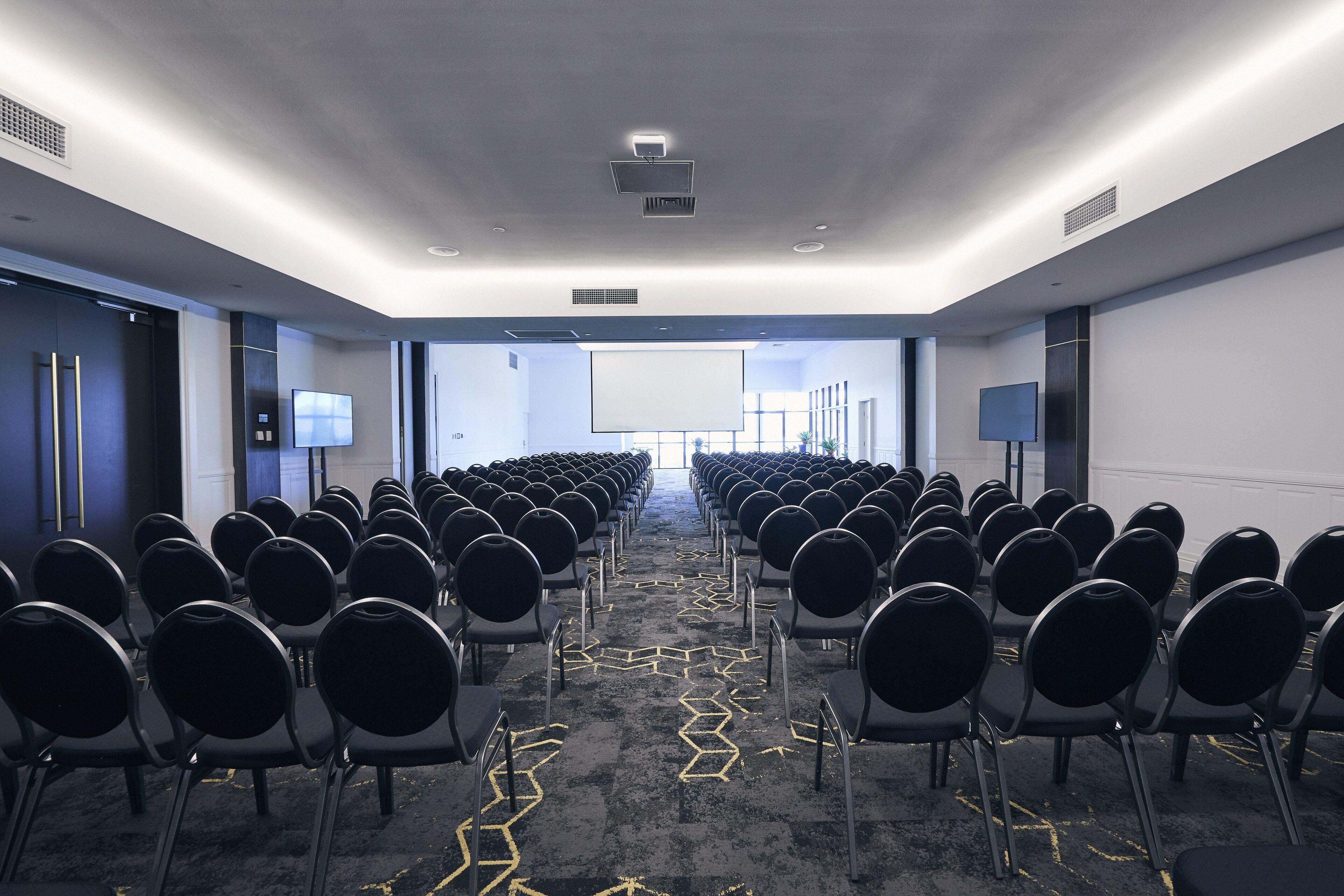 Un salón de conferencias con filas de sillas negras, una pantalla de proyector al frente y una alfombra con un diseño geométrico en zigzag amarillo.