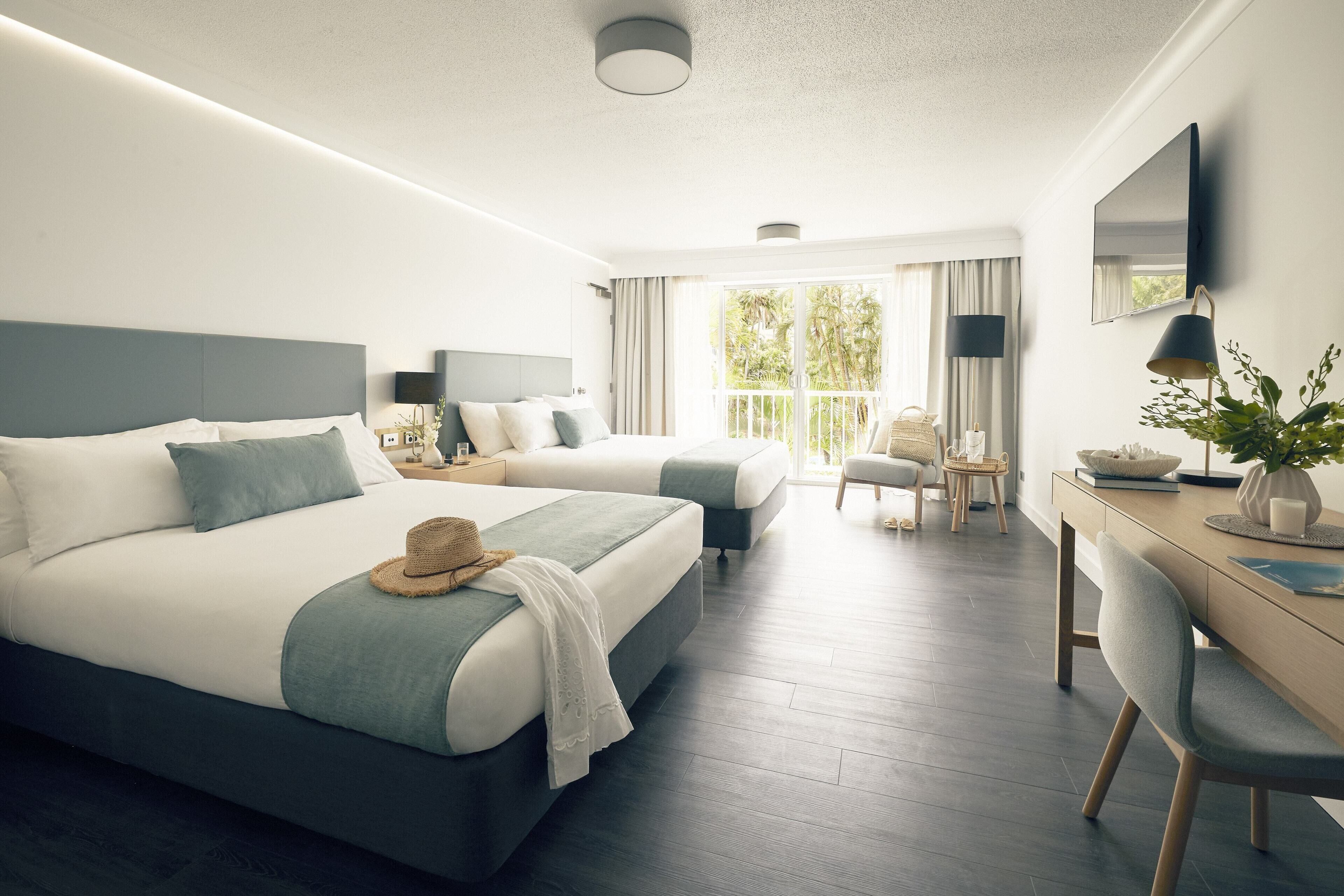 A hotel room with two beds, a desk, a chair, a TV, and a balcony with curtains.