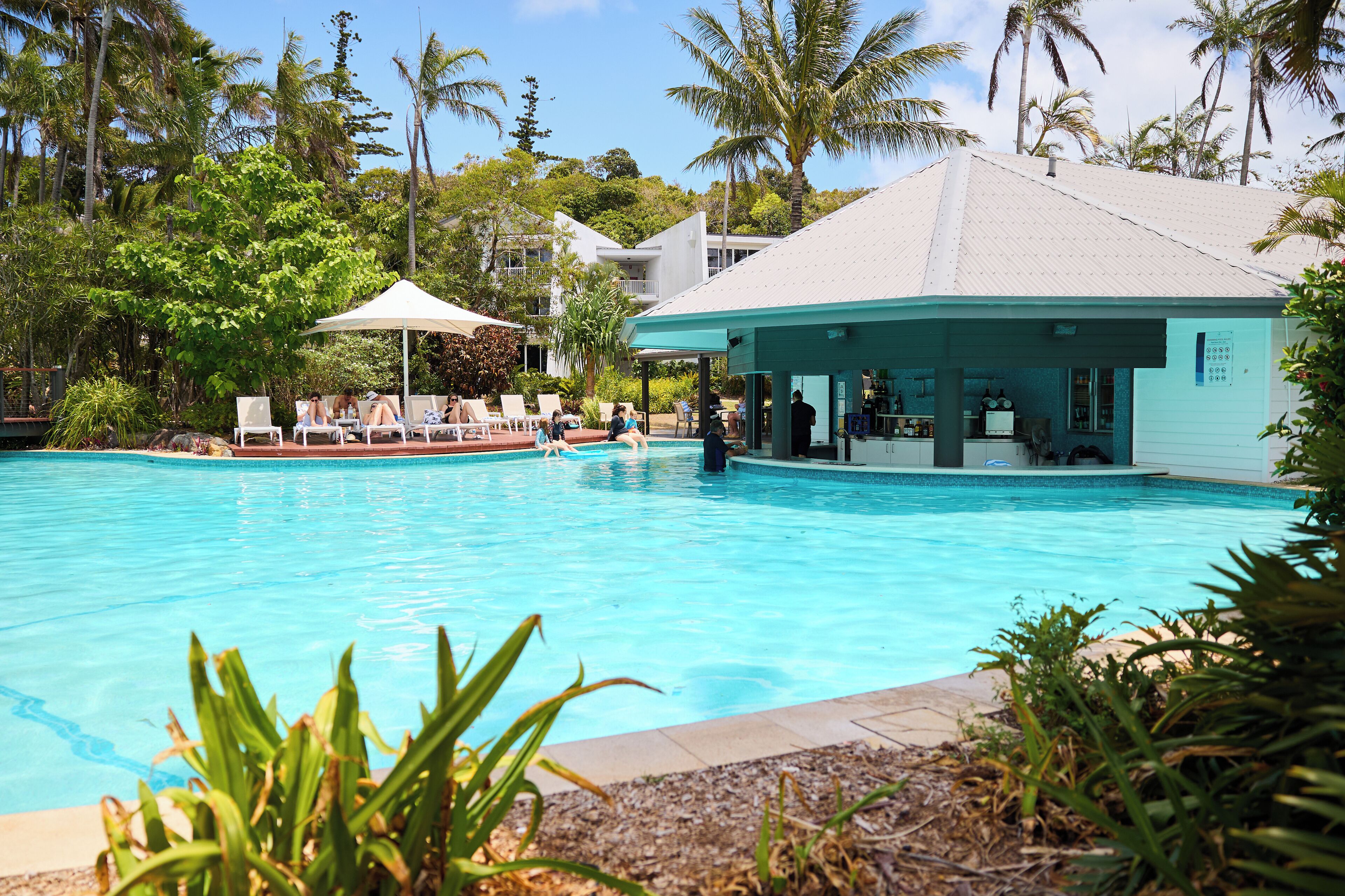 A swimming pool with lounge chairs and a covered area, surrounded by palm trees and a building with a white roof.