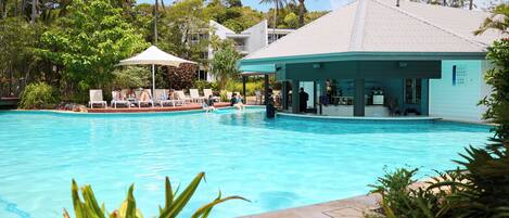A swimming pool with lounge chairs and a covered area, surrounded by palm trees and a building with a white roof.
