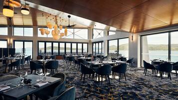 A spacious dining area with multiple tables and chairs, large windows offering views of the outdoors, and a ceiling with hanging lights.