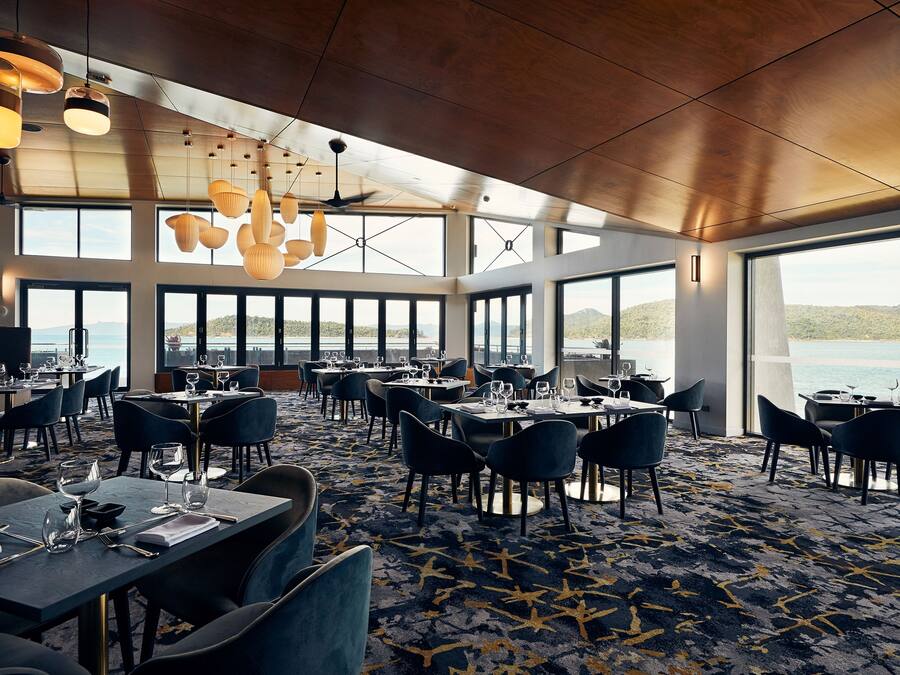 A spacious dining area with multiple tables and chairs, large windows offering views of the outdoors, and a ceiling with hanging lights.