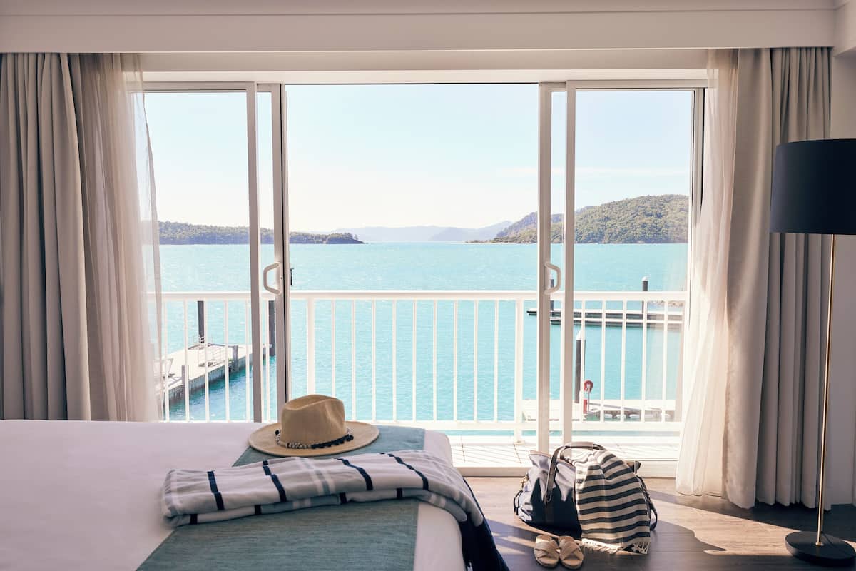 Habitación de hotel con un amplio ventanal corredera que da a un balcón con vistas a un lago y colinas al fondo.