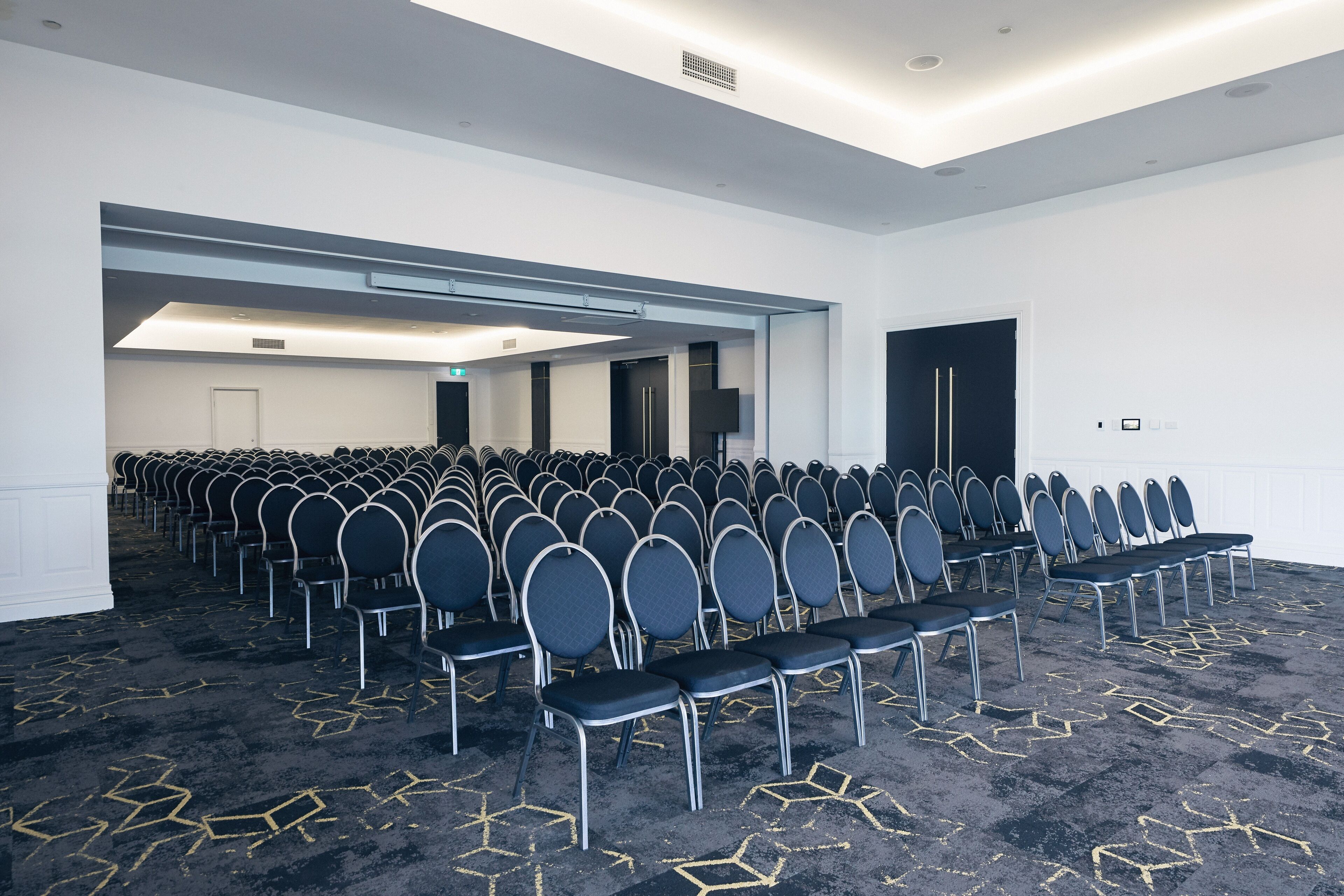 A conference room with rows of chairs, a carpeted floor, and a white wall with a door and a light fixture.