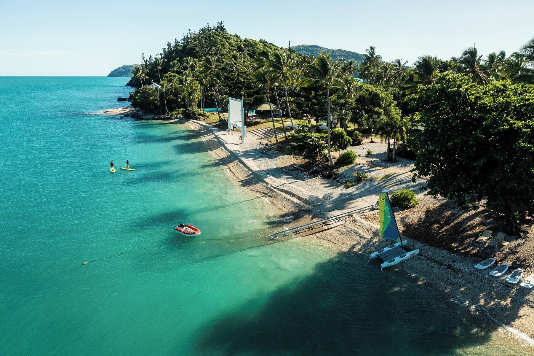 An aerial view of a beach with clear turquoise waters, a sandy shore, and dense tropical vegetation.
