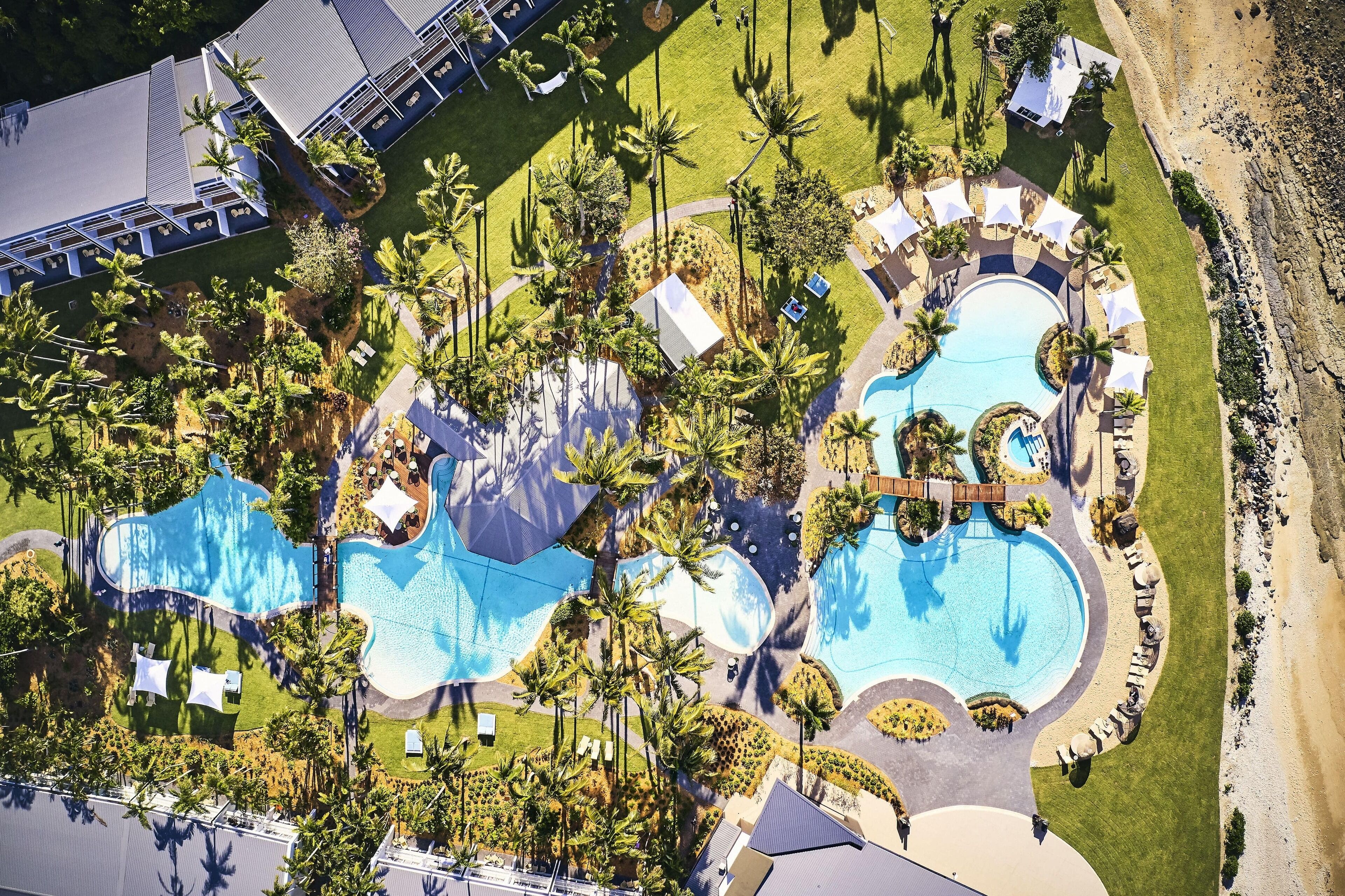 An aerial view of a resort with multiple swimming pools, palm trees, and a beach area.