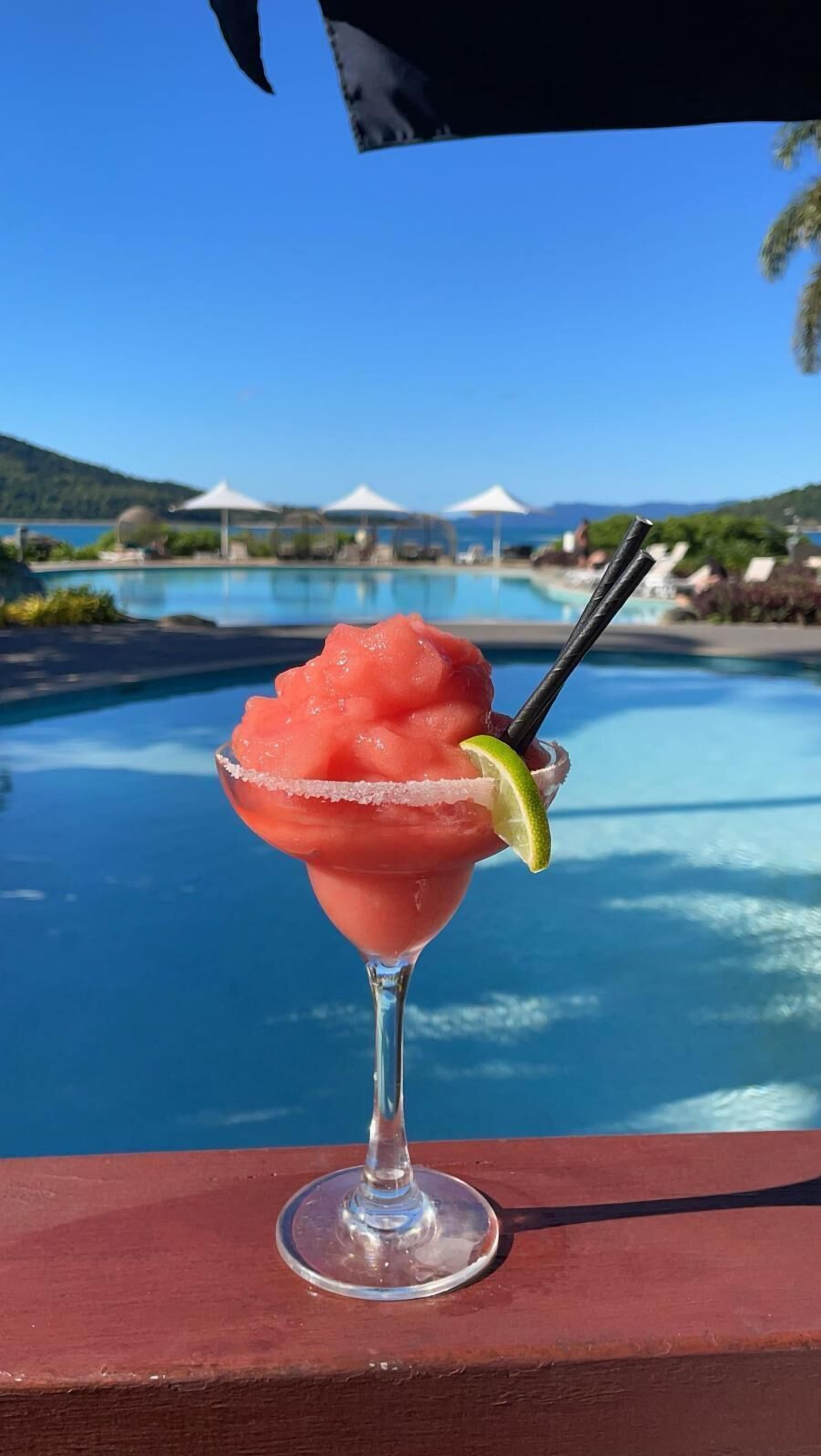 Ein rosa Cocktail mit Limettenscheibe und Salzrand, platziert auf einer Holzterrasse mit Blick auf einen Pool, der Sonnenschirme und Liegestühle hat.